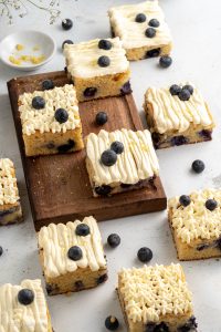 Squares of Keto Blueberry Lemon Cake with No Sugar Cream Cheese Frosting on wood board scattered on counter